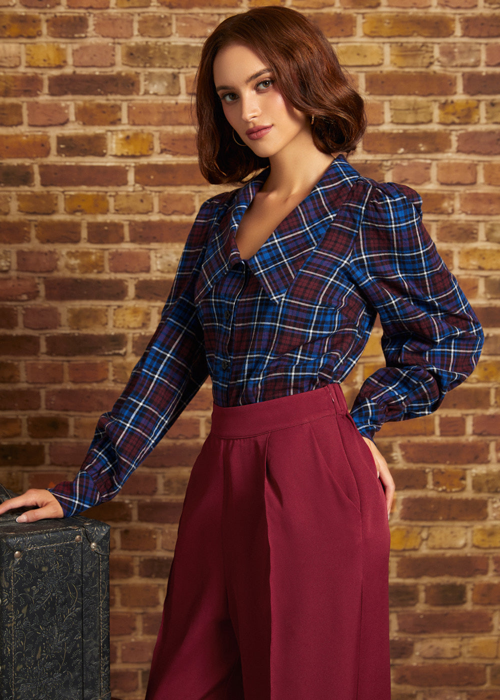 Model wearing a vintage-inspired burgundy and blue plaid blouse tucked into high-waisted burgundy trousers, showing its dramatic collar and softly puffed long sleeves.