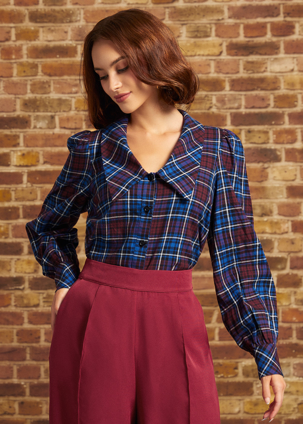 A woman wearing a blue and red plaid button-up blouse with puff sleeves and a high collar, paired with a high-waisted burgundy pants from ModCloth