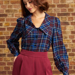 A woman wearing a blue and red plaid button-up blouse with puff sleeves and a high collar, paired with a high-waisted burgundy pants from ModCloth