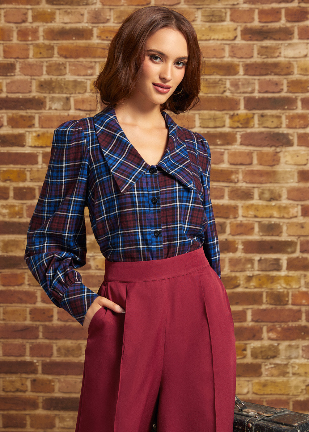 A woman wearing a blue and red plaid button-up shirt with a wide collar and long sleeves, paired with high-waisted burgundy wide-leg trousers, standing in front of a brick wall.