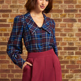 A woman wearing a blue and red plaid button-up shirt with a wide collar and long sleeves, paired with high-waisted burgundy wide-leg trousers, standing in front of a brick wall.