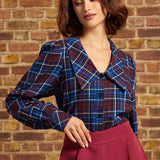 A woman wearing a blue and burgundy plaid button-up blouse with a large collar, paired with a high-waisted burgundy skirt, standing against a rustic brick wall.