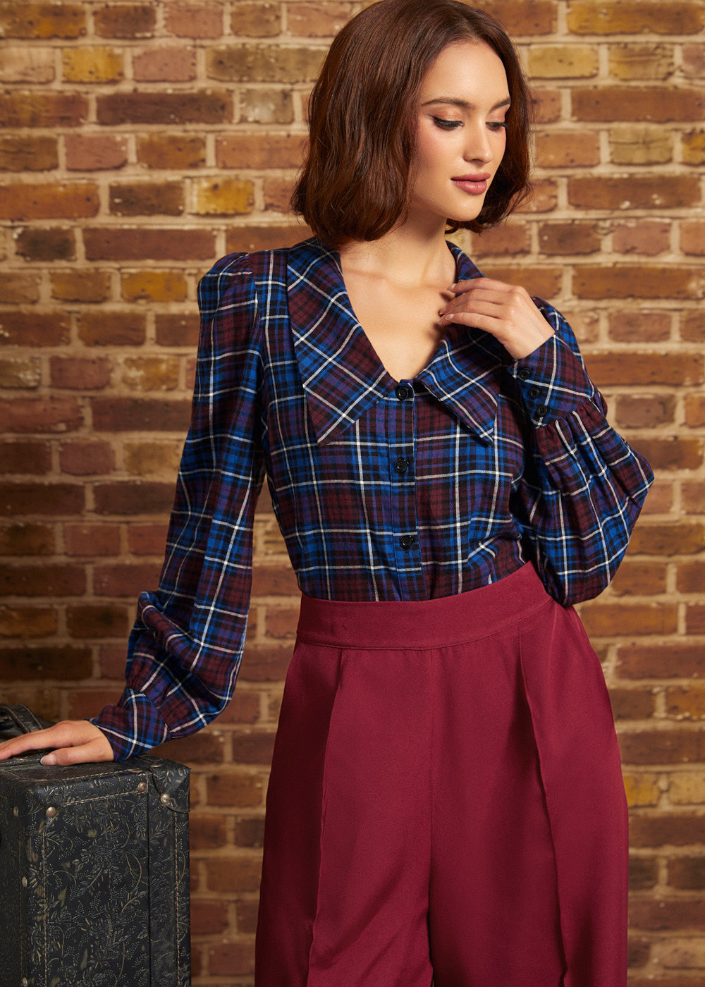 A woman wearing a blue and red plaid long-sleeve blouse with puffed shoulders and a buttoned V-neck, paired with high-waisted burgundy trousers, standing against a brick wall.