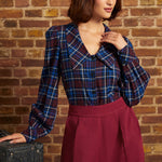 A woman wearing a blue and red plaid long-sleeve blouse with puffed shoulders and a buttoned V-neck, paired with high-waisted burgundy trousers, standing against a brick wall.