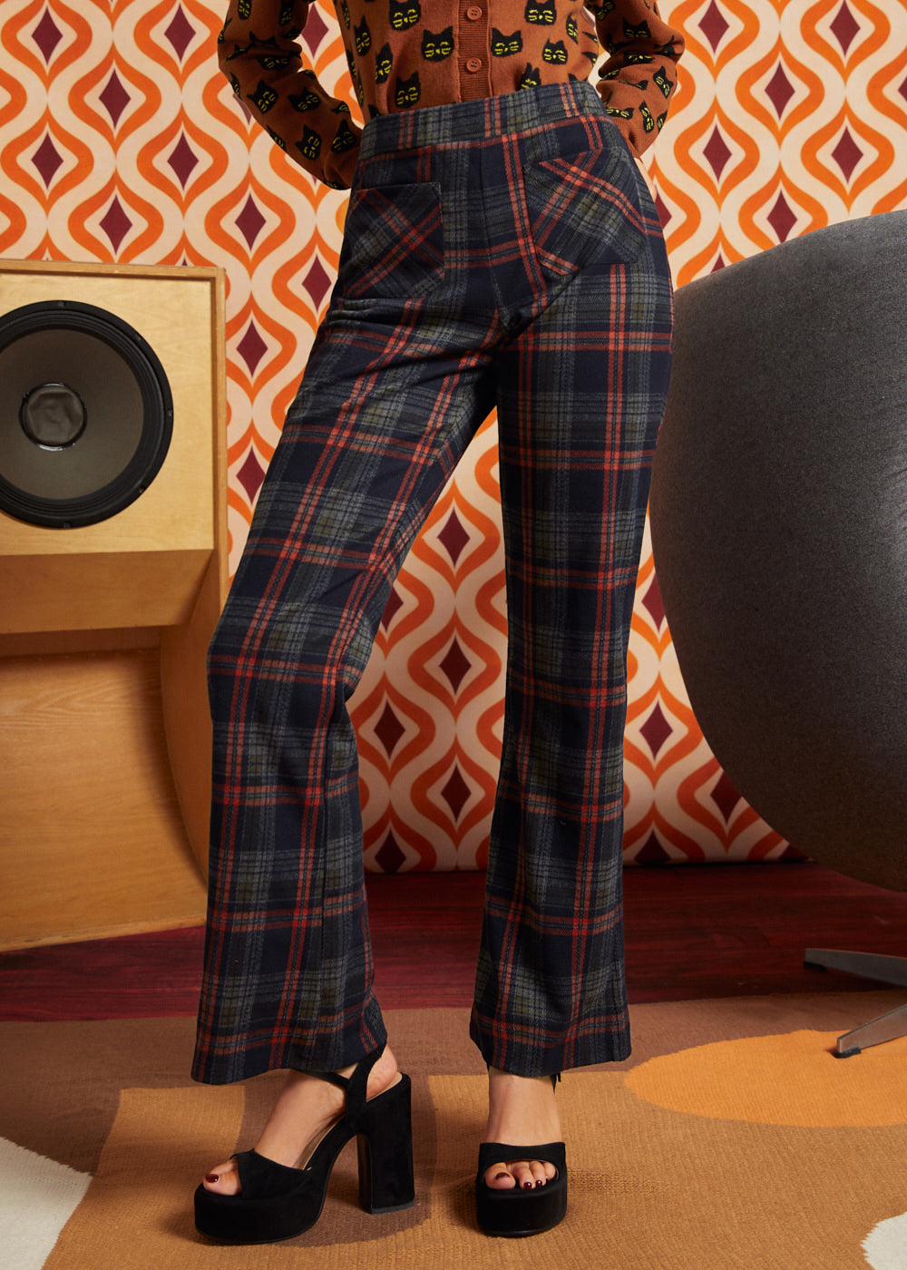 Woman wearing dark navy plaid high-waisted pants with rust and olive accents, brown cat cardigan, and black platform heels, front view