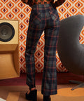 A woman shown from the back wearing high-waisted navy plaid trousers with red and orange lines, paired with a patterned top and black heels, standing in a retro-styled room with a speaker and geometric wallpaper.