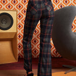 A woman shown from the back wearing high-waisted navy plaid trousers with red and orange lines, paired with a patterned top and black heels, standing in a retro-styled room with a speaker and geometric wallpaper.