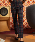 A woman wearing high-waisted dark plaid trousers with red, blue, and gray lines, paired with black platform sandals, standing in a retro-styled room with patterned wallpaper, a speaker, and a chair.