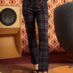 A woman wearing high-waisted dark plaid trousers with red, blue, and gray lines, paired with black platform sandals, standing in a retro-styled room with patterned wallpaper, a speaker, and a chair.