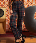 A model wearing high-waisted, wide-leg plaid trousers in navy, red, and black, paired with a brown cat-print top and black platform heels, standing in a retro-styled room with a patterned orange and white wallpaper background.