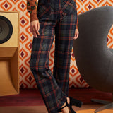 A model wearing high-waisted, wide-leg plaid trousers in navy, red, and black, paired with a brown cat-print top and black platform heels, standing in a retro-styled room with a patterned orange and white wallpaper background.