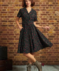 A woman models a short-sleeve black dress with a red and green floral print, featuring a collared neckline, button-front bodice, and a flared skirt. She is standing in front of a rustic brick wall, wearing brown lace-up ankle boots.