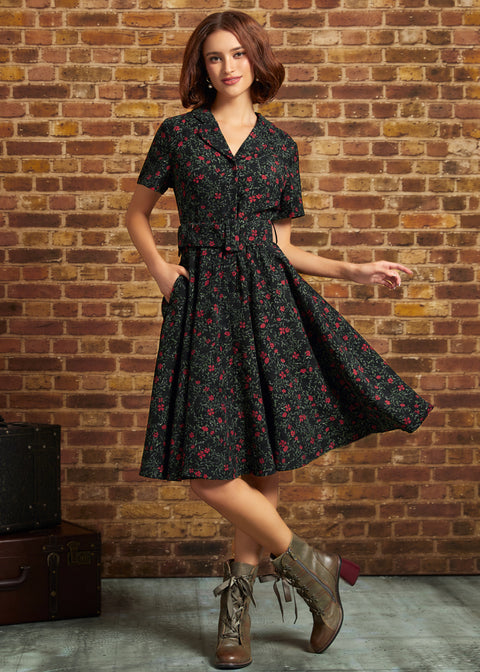 A woman models a short-sleeve black dress with a red and green floral print, featuring a collared neckline, button-front bodice, and a flared skirt. She is standing in front of a rustic brick wall, wearing brown lace-up ankle boots.