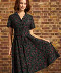 Model wearing a black floral corduroy midi dress with red roses, short sleeves, notched collar, button-front bodice, and matching buckled belt against a brick wall.