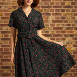 Model wearing a black floral corduroy midi dress with red roses, short sleeves, notched collar, button-front bodice, and matching buckled belt against a brick wall.