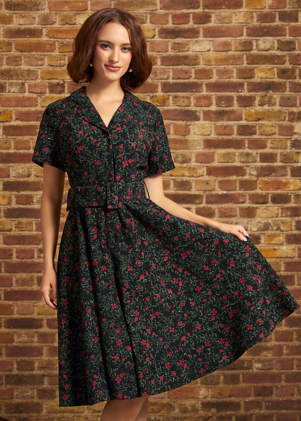 Model wearing a black floral corduroy midi dress with red roses, short sleeves, notched collar, button-front bodice, and matching buckled belt against a brick wall.