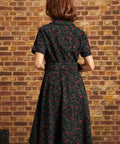 Back view of a model wearing a black floral corduroy midi dress with short sleeves, a fitted waist, matching belt, and full skirt against a brick wall.