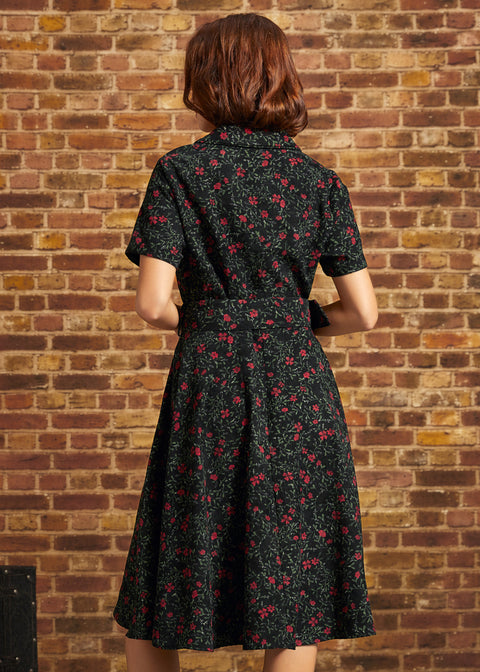 Back view of a model wearing a black floral corduroy midi dress with short sleeves, a fitted waist, matching belt, and full skirt against a brick wall.