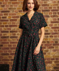 A woman wearing a short-sleeve black ModCloth dress with a red and pink floral print, standing in front of a rustic brick wall.