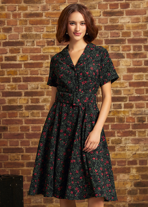 A woman wearing a short-sleeve black ModCloth dress with a red and pink floral print, standing in front of a rustic brick wall.