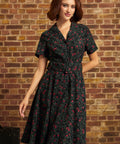 A woman wearing a short-sleeve black shirt dress with a red and green floral print, standing in front of a brick wall.