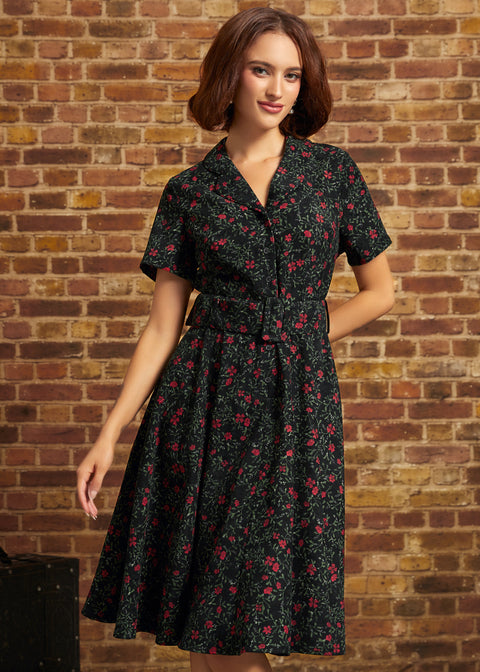 A woman wearing a short-sleeve black shirt dress with a red and green floral print, standing in front of a brick wall.