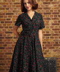 A woman wearing a short-sleeve black dress with a red and pink floral print, standing in front of a brick wall next to a vintage trunk.