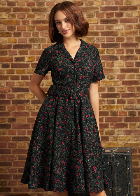 A woman wearing a short-sleeve black dress with a red and pink floral print, standing in front of a brick wall next to a vintage trunk.