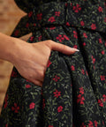 Close-up of a hand tucked into the side seam pocket of a black corduroy dress featuring small red flowers and green botanical vines.