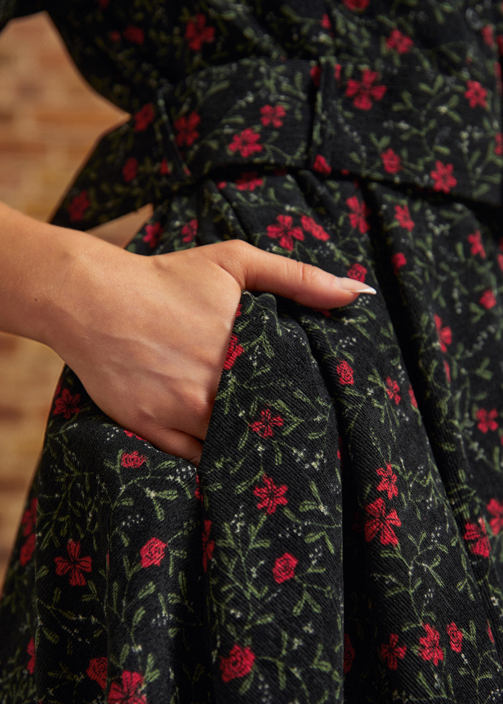 Close-up of a hand tucked into the side seam pocket of a black corduroy dress featuring small red flowers and green botanical vines.