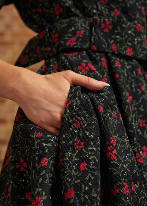 Close-up of a hand tucked into the side seam pocket of a black corduroy dress featuring small red flowers and green botanical vines.