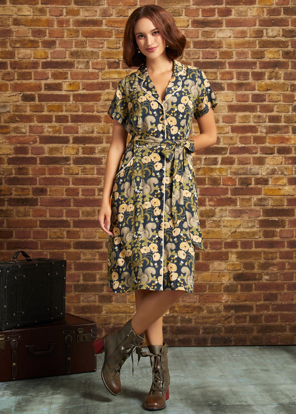 A woman models a short-sleeve floral shirt dress with a tie waist, standing in front of a brick wall. She is wearing brown lace-up boots, and a vintage suitcase sits on the floor to her left.