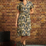 A woman models a short-sleeve floral shirt dress with a tie waist, standing in front of a brick wall. She is wearing brown lace-up boots, and a vintage suitcase sits on the floor to her left.
