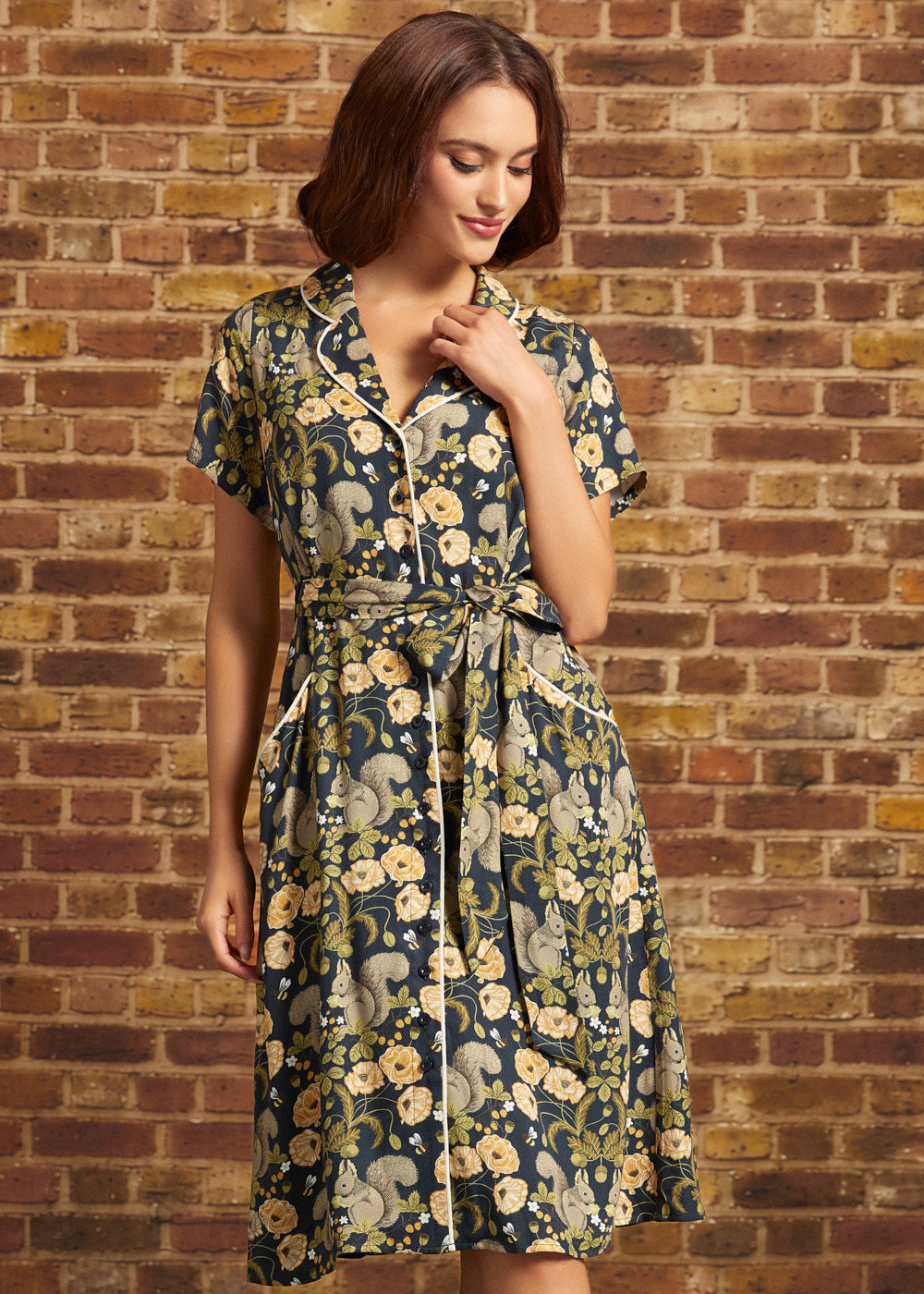 Model wearing a vintage-inspired navy squirrel and floral print button-front midi dress with short sleeves, contrast piping, and a tied waist belt.