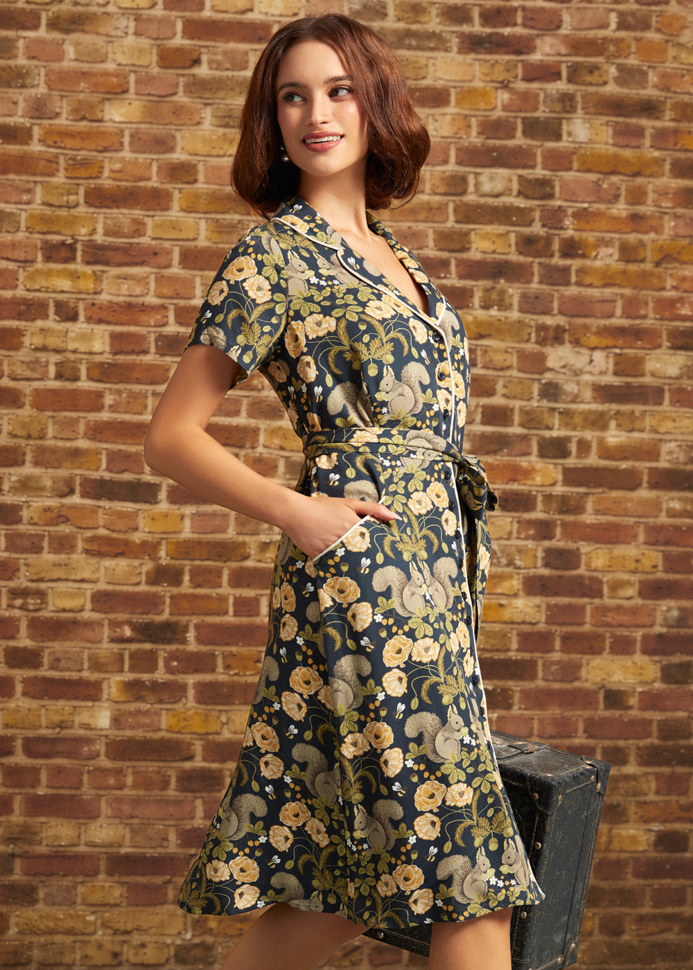 A woman models a short-sleeve shirt dress with a navy blue background and a beige and yellow floral and squirrel pattern, standing against a brick wall.