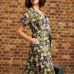 A woman models a short-sleeve shirt dress with a navy blue background and a beige and yellow floral and squirrel pattern, standing against a brick wall.