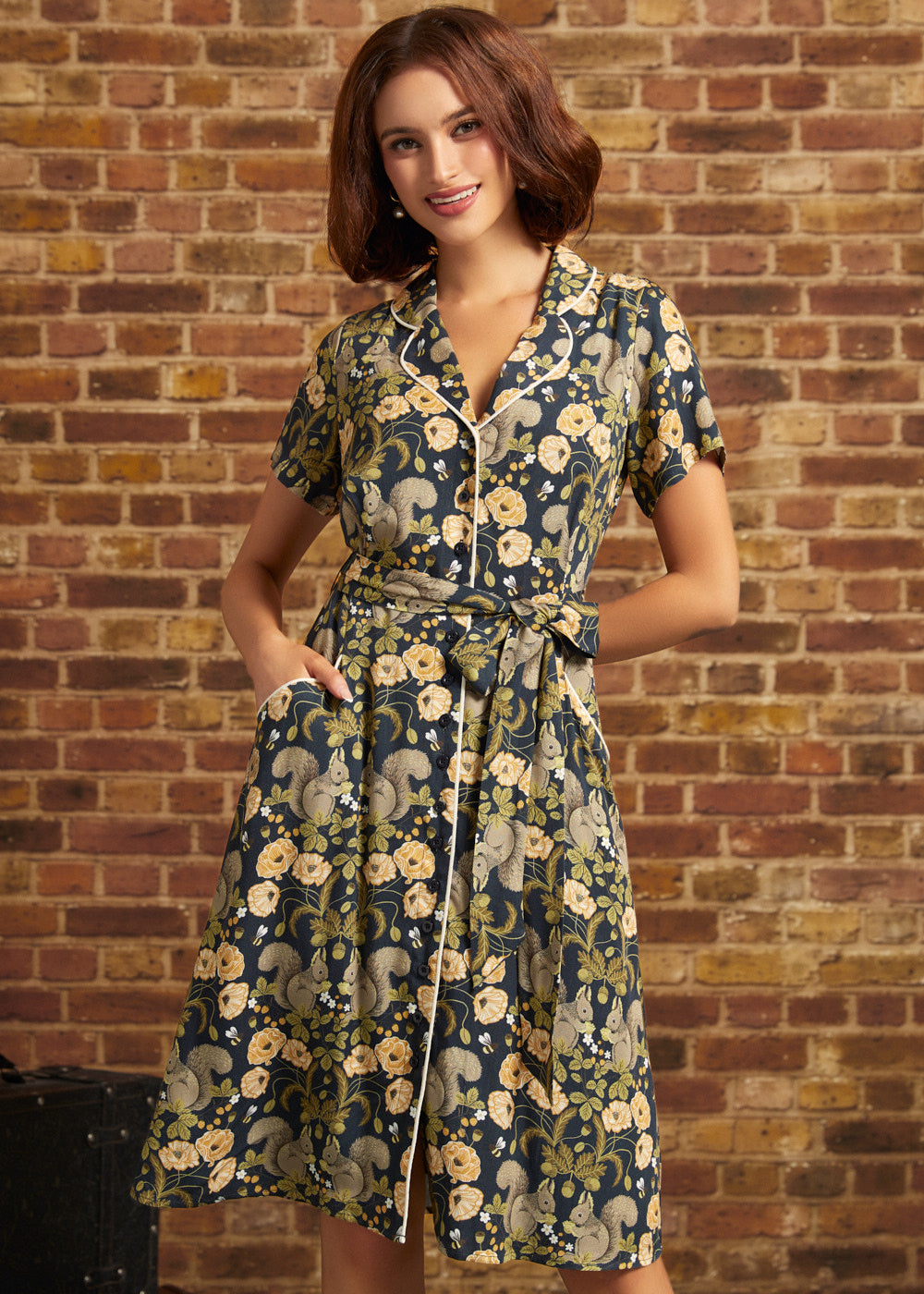 A woman wearing a short-sleeve button-front dress with a waist tie and pockets, featuring a navy blue and beige floral and squirrel print, standing in front of a brick wall.