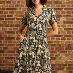 A woman wearing a short-sleeve button-front dress with a waist tie and pockets, featuring a navy blue and beige floral and squirrel print, standing in front of a brick wall.
