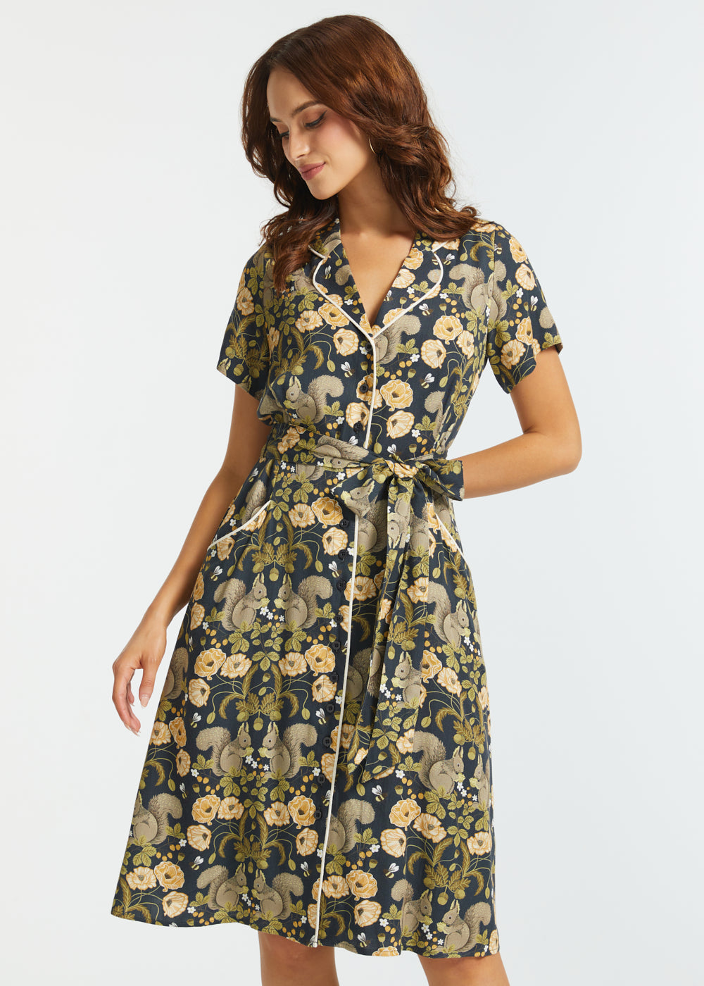 A woman modeling a short-sleeve midi dress with a dark navy background and a detailed floral and squirrel pattern in beige, olive green, and cream, set against a plain light gray studio background.