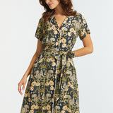 A woman modeling a short-sleeve midi dress with a dark navy background and a detailed floral and squirrel pattern in beige, olive green, and cream, set against a plain light gray studio background.