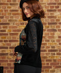 Side view of a woman wearing a ModCloth black lace long sleeve blouse with a cat print tapestry vest on top