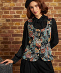 A woman wearing a black lace long-sleeve blouse under a colorful floral-patterned vest, paired with black trousers and a black beret, posing against a brick wall.