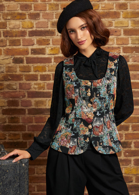 A woman wearing a black lace long-sleeve blouse under a colorful floral-patterned vest, paired with black trousers and a black beret, posing against a brick wall.