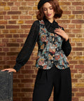 A woman wearing a black beret, a black sheer long-sleeve blouse, and a black vest with a colorful floral print, paired with black wide-leg trousers, posing in front of a brick wall.