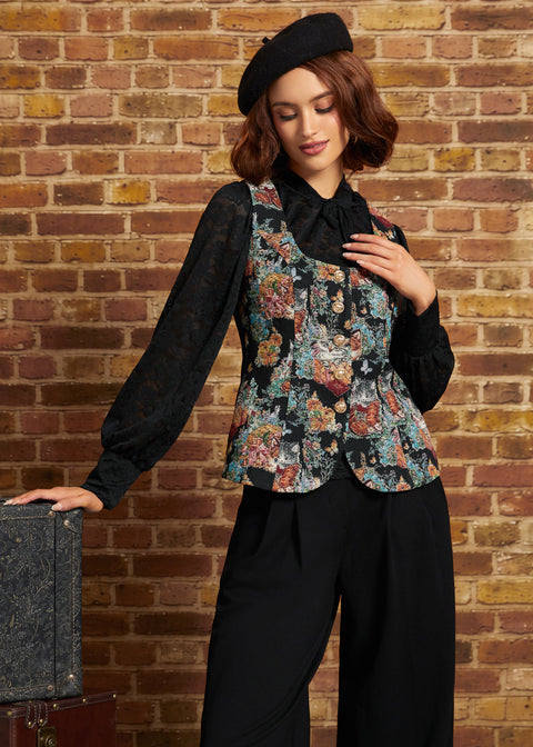 A woman wearing a black beret, a black sheer long-sleeve blouse, and a black vest with a colorful floral print, paired with black wide-leg trousers, posing in front of a brick wall.