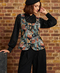 A woman wearing a black lace long-sleeve blouse under a black vest with a colorful floral print, black wide-leg pants, and a black beret, posing in front of a brick wall.