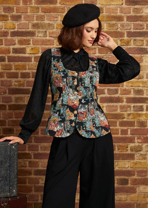 A woman wearing a black lace long-sleeve blouse under a black vest with a colorful floral print, black wide-leg pants, and a black beret, posing in front of a brick wall.