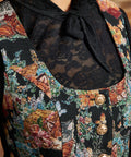 Close-up of the ModCloth Winifred Waistcoat Vest showing its scoop neckline, colorful woven-style cat and floral design, and decorative gold-tone buttons, layered over a black lace bow blouse.