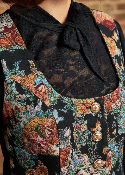Close-up of the ModCloth Winifred Waistcoat Vest showing its scoop neckline, colorful woven-style cat and floral design, and decorative gold-tone buttons, layered over a black lace bow blouse.
