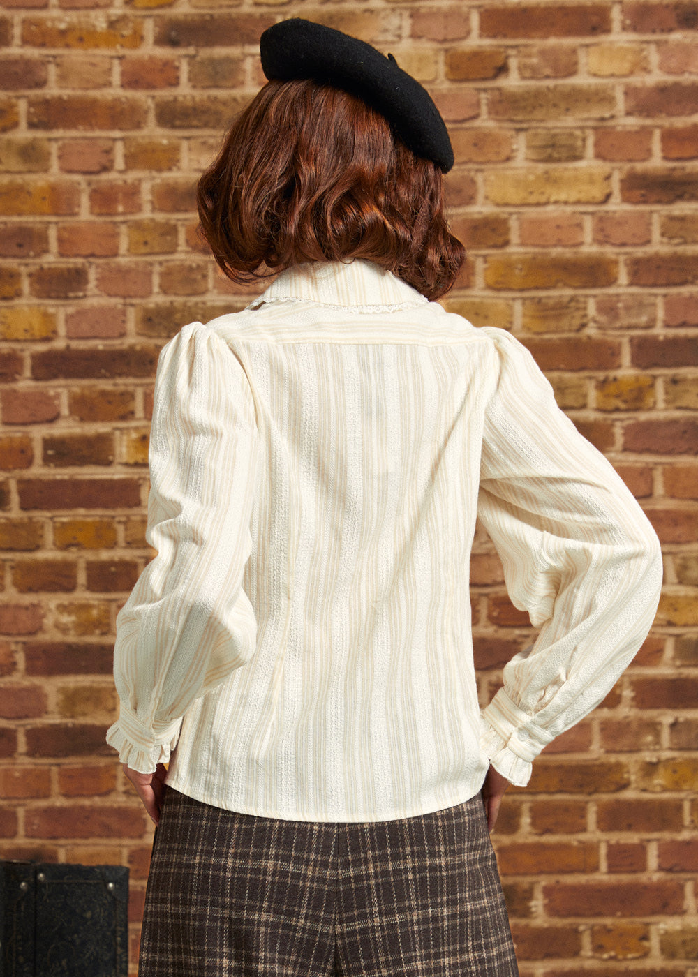 Back view of an ivory Victorian-inspired blouse with textured vertical stripes, long puff sleeves, and ruffled cuffs, styled with plaid trousers and a black beret.